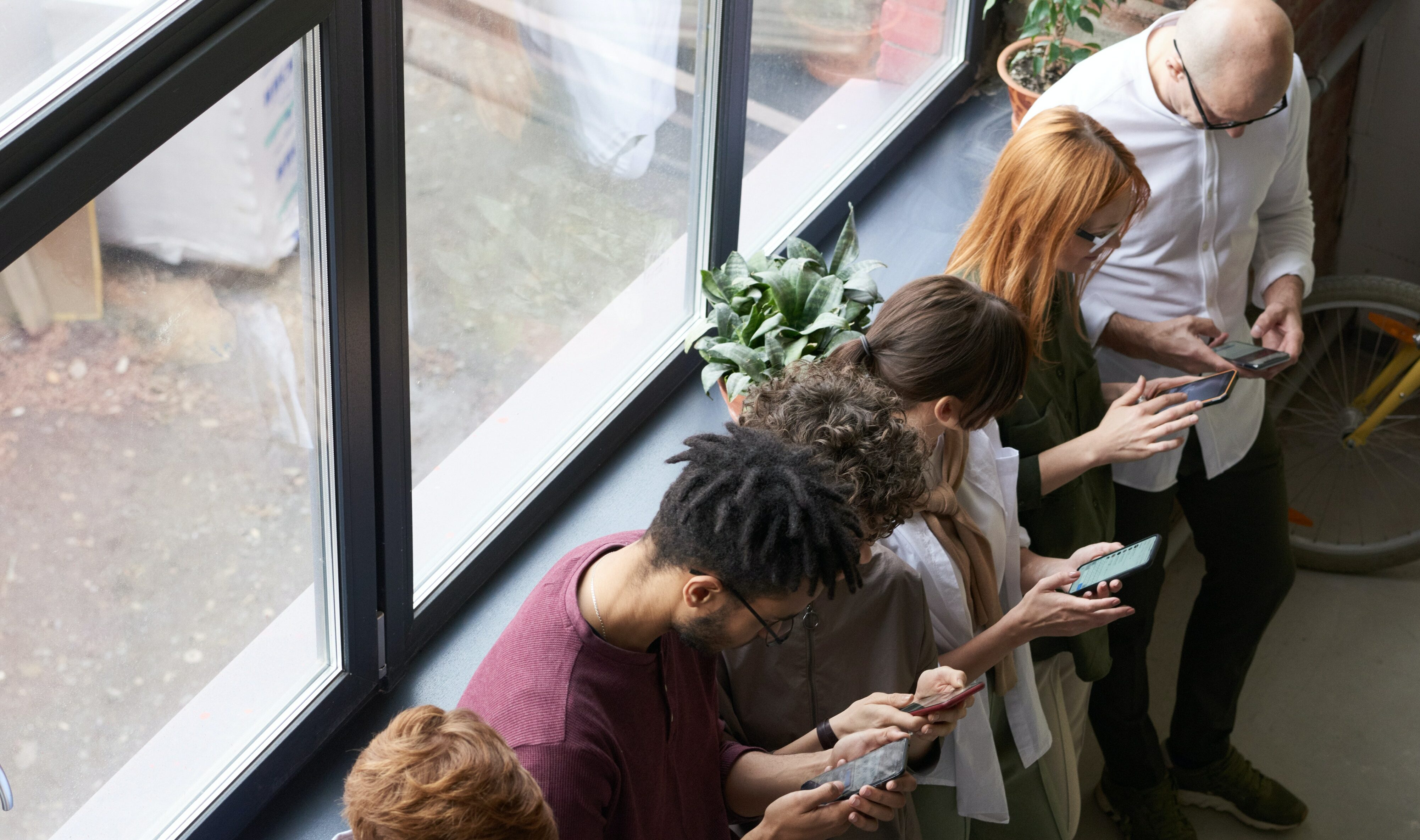 Eight people stand in a line leaning on a windowsill with their heads in their phones on social media