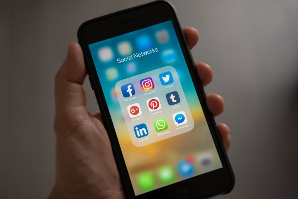 A hand holding a smart phone with social networking icons showing Facebook, Instagram, Twitter, Pinterest, Twitter, LinkedIn, WhatsApp, and Facebook Messenger