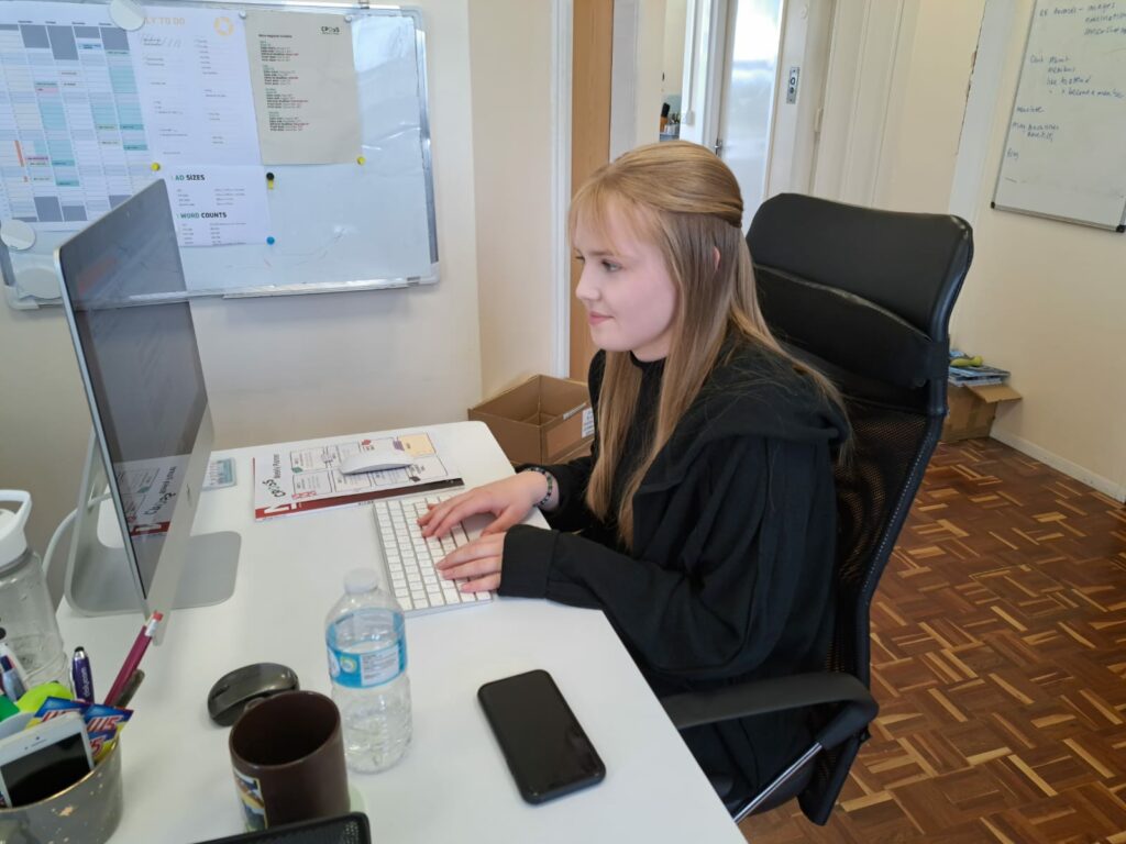 Work experience student Alice Kotulecki sits at a white desk smiling at a Mac computer