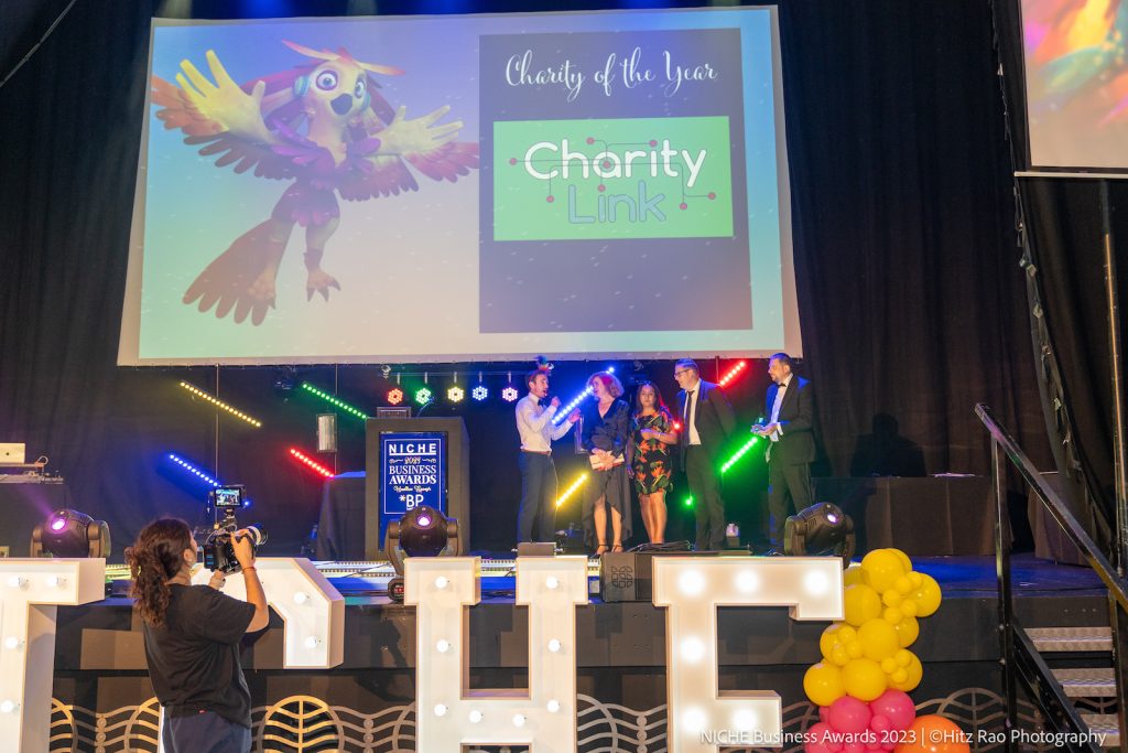 A business event stage with a compere giving an award to Charity Link of Leicestershire at the Niche Business Awards 2023